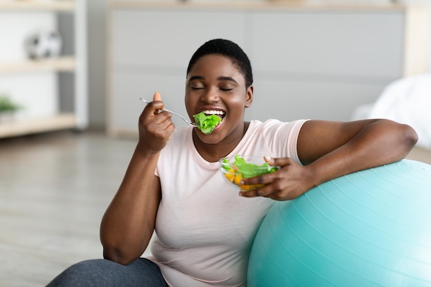 Sport en gezond eten voor afslankende mollige zwarte vrouw met fitnessbal die geniet van smakelijke groente