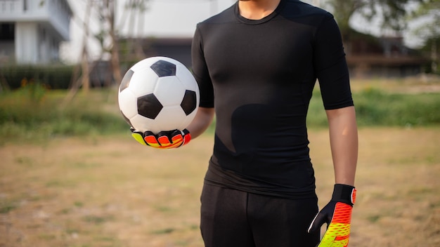 Sport- en recreatieconcept een mannelijke tienerkeeper die een zwarte outfit draagt en een paar kleurrijke handschoenen met een voetbal.