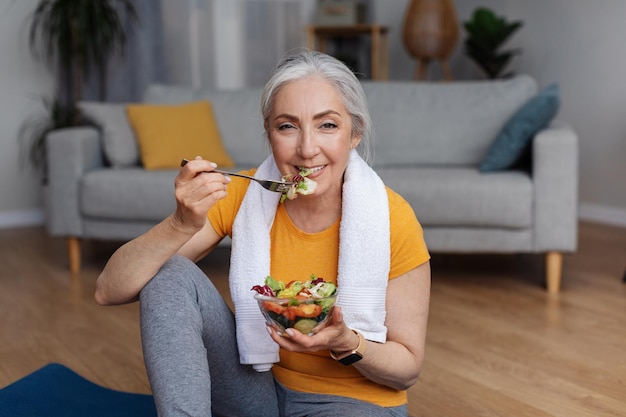 Sport- en voedingsconcept Happy senior femael leunend met het eten van verse groentesalade die thuis op yogamat zit