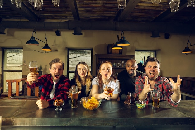 Sport fans cheering at bar, pub and drinking beer while championship, competition is going. Multiethnic group of friends excited watching translation. Human emotions, expression, supporting concept.