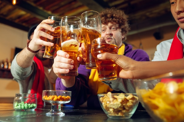 Sport fans cheering at bar, pub and drinking beer while championship, competition is going. Multiethnic group of friends excited watching translation. Human emotions, expression, supporting concept.