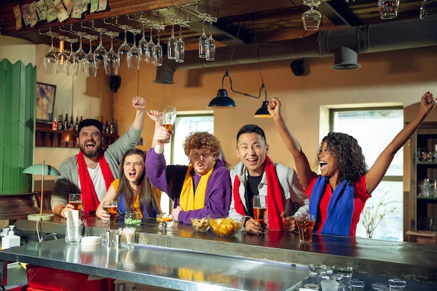 Sport fans cheering at bar, pub and drinking beer while championship, competition is going. Multiethnic group of friends.