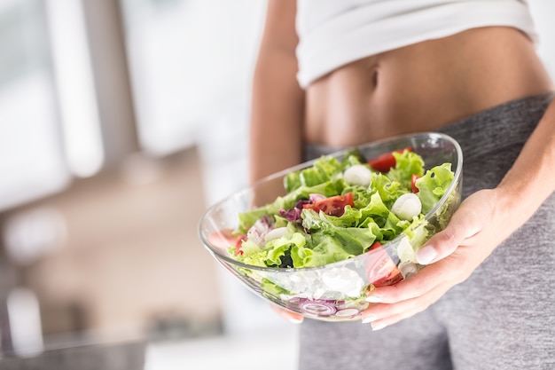 La figura sportiva di una donna attraente che tiene in mano una ciotola con insalata di verdure fresche.