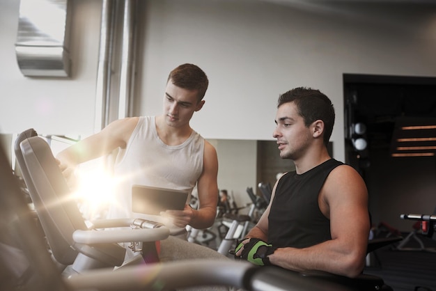 sport, fitness, equipment, technology and people concept - men with tablet pc computer exercising on gym machine