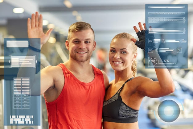 sport, fitness, gesture and people concept - smiling man and woman waving hands in gym over virtual charts