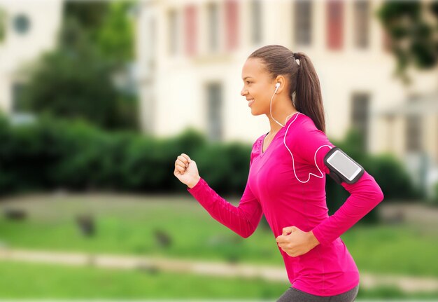 sport, fitness, gezondheid, technologie en mensenconcept - glimlachende jonge Afro-Amerikaanse vrouw die buiten met smartphone en oortelefoons loopt