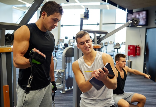 sport, fitness, lifestyle and people concept - men with clipboard taking notes and exercising on gym machine