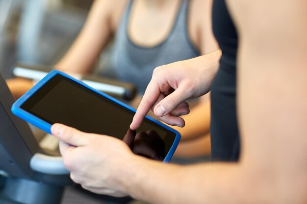 sport, fitness, lifestyle, technology and people concept - close up of trainer hands with tablet pc computer and woman working out on exercise bike in gym