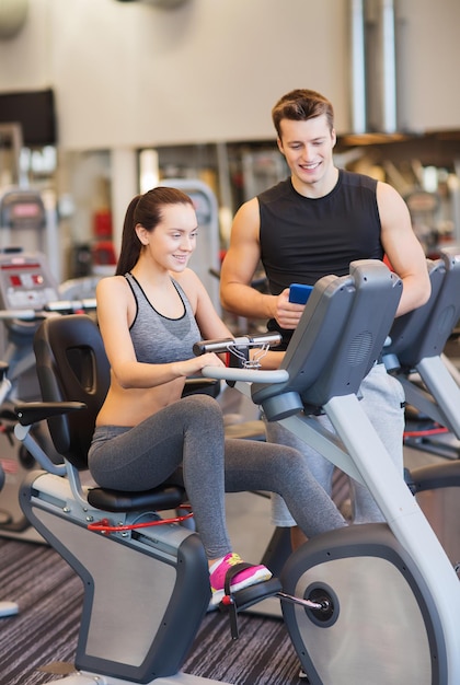 sport, fitness, lifestyle, technology and people concept - happy woman with trainer working out on exercise bike in gym