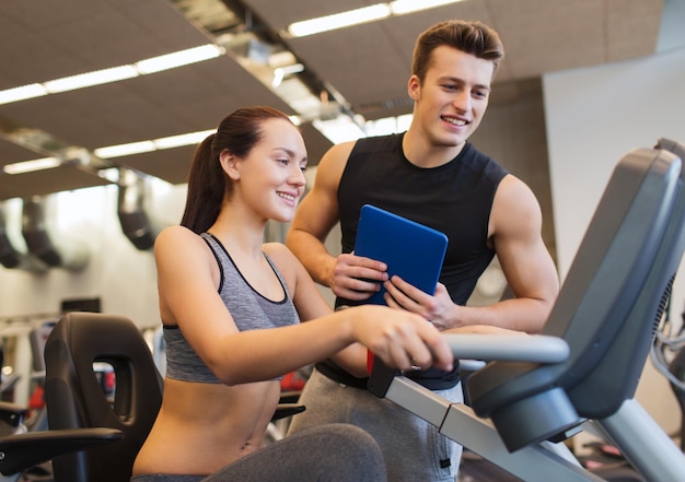 Sport, fitness, stile di vita, tecnologia e concetto di persone - donna felice con trainer che si allena sulla cyclette in palestra