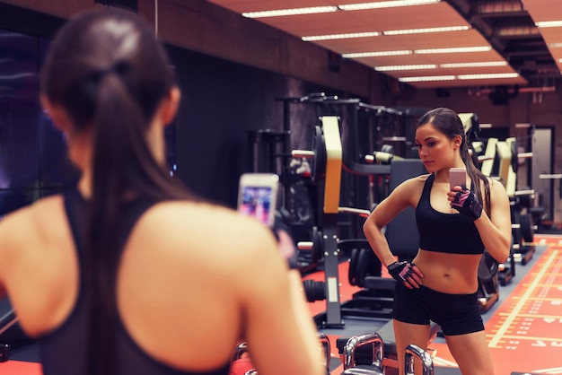 спорт, фитнес, образ жизни, технологии и концепция людей - молодая женщина со смартфоном делает селфи в зеркале в тренажерном зале