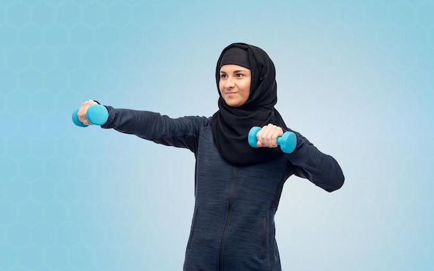 Photo sport, fitness and people concept - happy muslim woman in hijab with dumbbells doing exercises over blue background