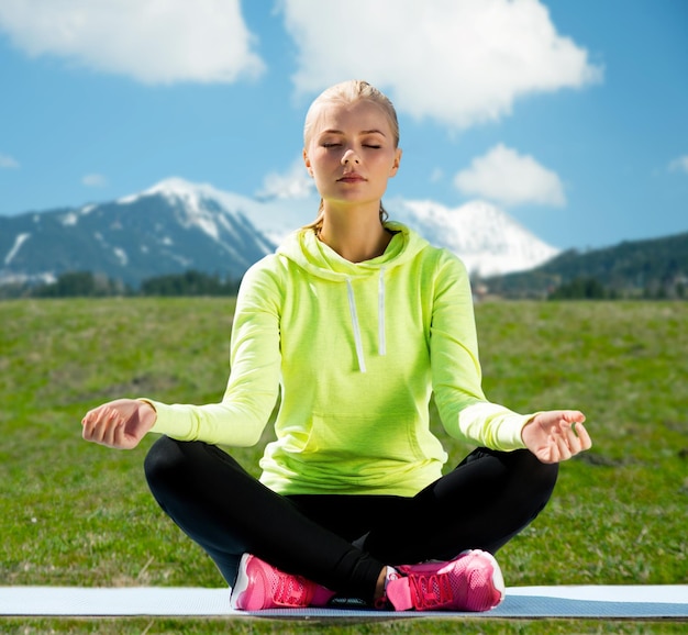 sport, fitness, people and meditation concept - woman sitting in lotus pose doing yoga over mountains, green field and blue sky background