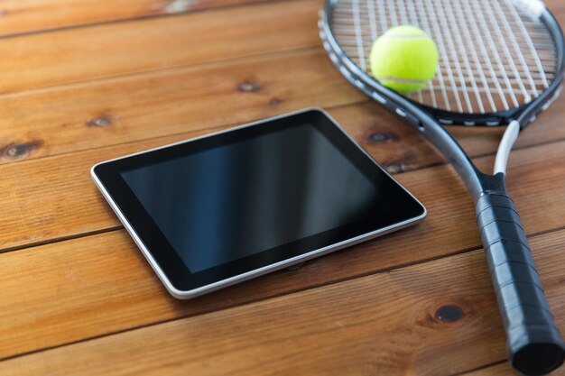 sport, fitness, technology, game and objects concept - close up of tennis racket with ball and tablet pc computer on wooden floor