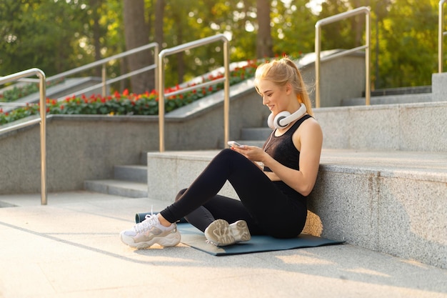 スポーツとフィットネス若い大人の白人女性スマートフォンを使用して座っているエクササイズマットコンクリートの床朝のトレーニングの後に休む屋外サマーパークアスレチック女性休憩をとる携帯電話を使用する