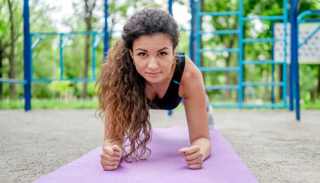 Спортивная девушка делает упражнения планки на циновке для йоги на открытом воздухе и смотрит в камеру. Молодая женщина во время тренировки на стадионе