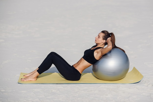Спорт и концепция здорового образа жизни. Красивая девушка фитнеса делая тренировки живота с подходящим шариком на пляже. Молодая женщина делает упражнения пилатес.