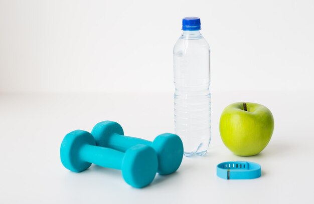 sport, healthy lifestyle and objects concept - close up of dumbbells with fitness tracker, green apple and water bottle over white background