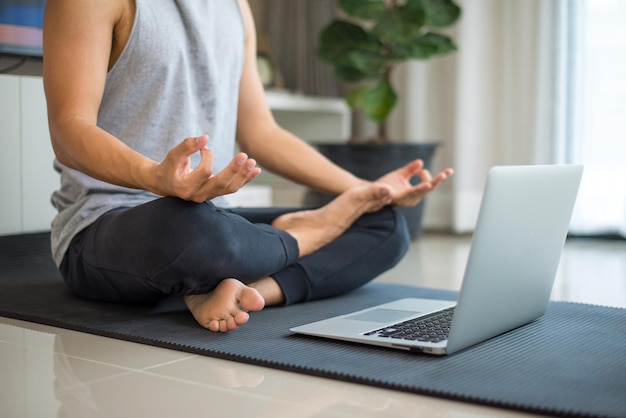 Uomo sportivo seduto sul tappetino per esercizi che pratica meditazione con istruttore di fitness che prende lezioni di yoga online durante una videochiamata nel laptop nel soggiorno di casa