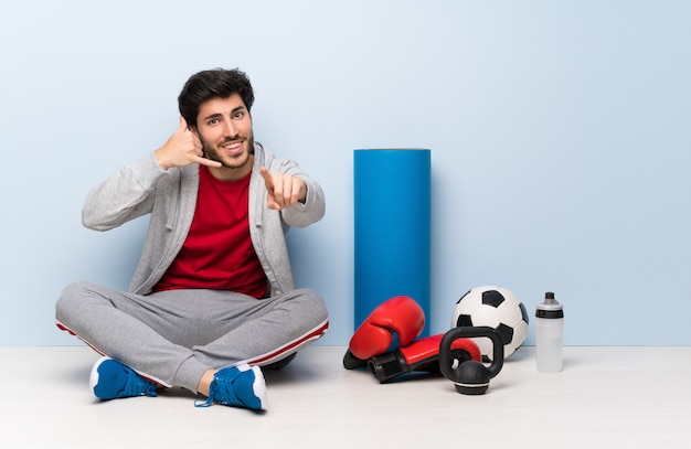 Sport man sitting on the floor making phone gesture and pointing front