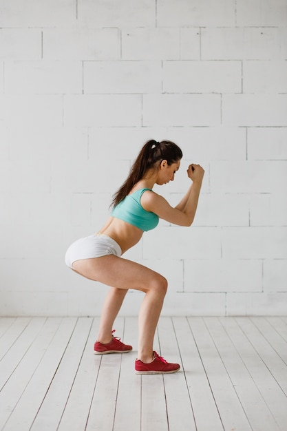 Sport meisje doet warming-up, squats. het mooie meisje leidt in witte studio op. ochtend gymnastiek. jonge vrouw gekleed in aquamarijn top, witte korte broek, rode sneackers