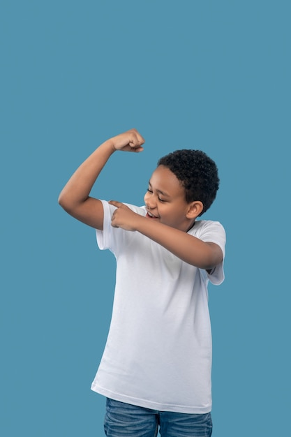 Sport, prestatie. Optimistische jongen met een donkere huid in een wit t-shirt die zijn hand aanraakt en zich verheugt in sterke spieren die op een blauwe achtergrond staan