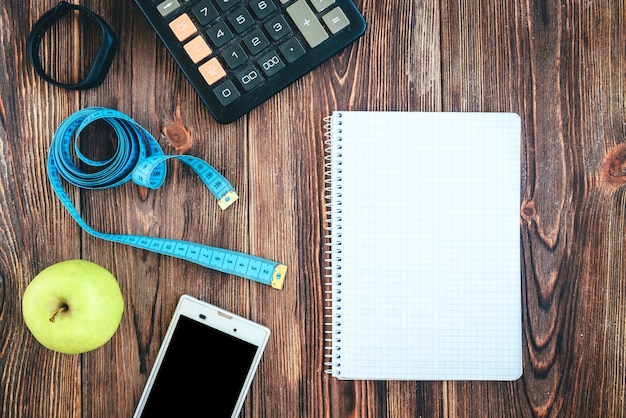 Sport shoes, smartphone, tape, calculator, fitness bracelet, green apple and notebook on wooden background.