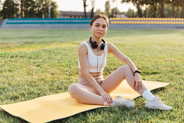 Photo sport with music. pleasant looking dark haired athlete woman wearing white top and beige leggins, with headphones over neck listening music while exercising in stadium.