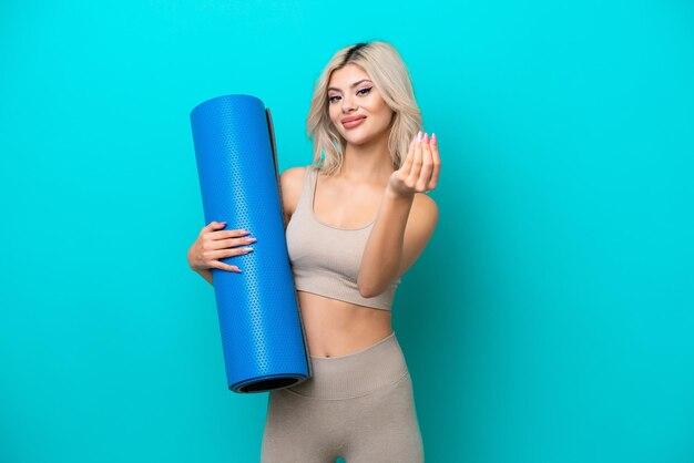 Sport woman going to yoga classes while holding a mat isolated on blue background making money gesture