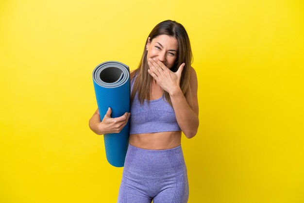 Sport woman going to yoga classes while holding a mat isolated non yellow background happy and smiling covering mouth with hand