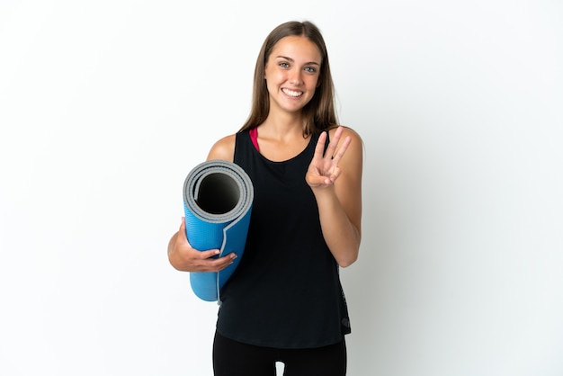 Sport woman going to yoga classes while holding a mat over isolated white background happy and counting three with fingers