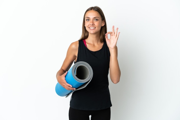 Donna sportiva che va alle lezioni di yoga mentre tiene in mano un tappetino su sfondo bianco isolato che mostra il segno ok con le dita