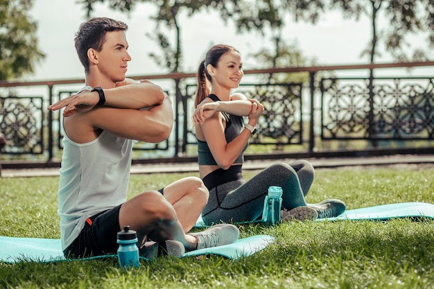 Sport woman and man warming together in the park while sitting on mats at daytime