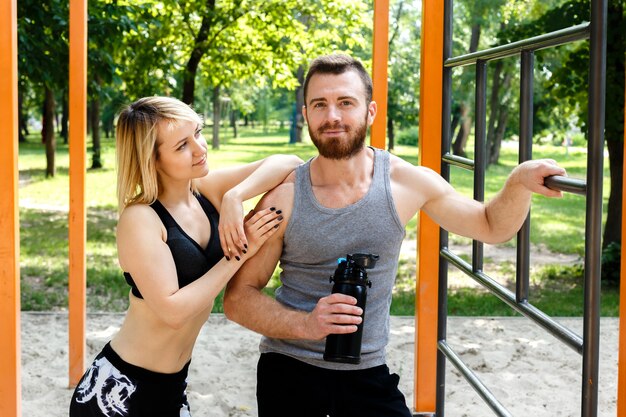 Sportief blondemeisje en gebaarde mens die na training opleiding rusten in een park openlucht. Mens die een zwarte fles met water houdt.