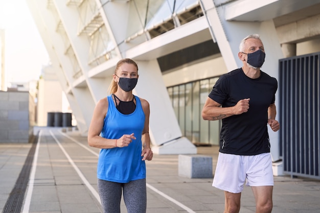 Sportief volwassen stel met beschermende gezichtsmaskers die samen joggen in de stedelijke omgeving