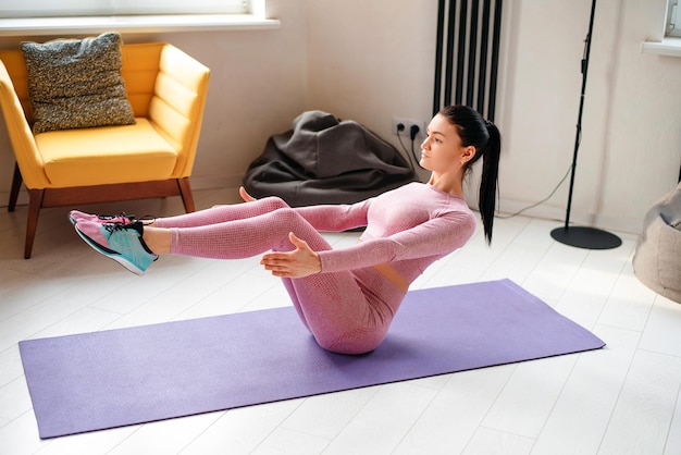 Sportieve blanke vrouw in sportkleding die buikspieren traint terwijl ze balanceert op yogamat Fitness jongedame met actieve training in modern appartement