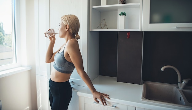 Sportieve blonde vrouw die activewear draagt, drinkt een glas water na fitness thuis