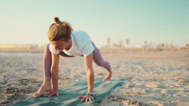 Foto sportieve blonde vrouw die yoga uitoefent op fitnessmat bij de zee aantrekkelijk yogi-meisje gekleed in sportkleding die ochtendoefeningen op het strand doet