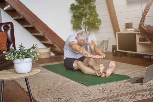 Sportieve, gezonde man van middelbare leeftijd die rekoefeningen doet terwijl hij op een yogamat in de woonkamer zit