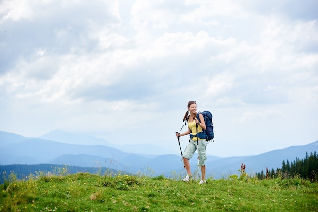 Sportieve glimlachende vrouw die backpacker in bergpad wandelen, op grasrijke heuvel lopen, trekkingsstokken gebruiken, die van de zomer bewolkte dag genieten. Outdoor-activiteit, lifestyle concept