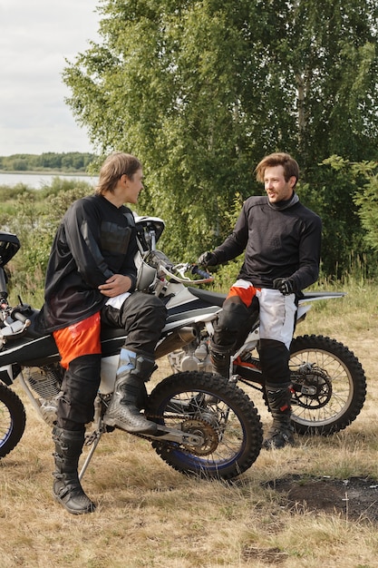 Sportieve jonge mannen in motoroutfits zittend op motoren en praten tijdens het rusten na race in het bos