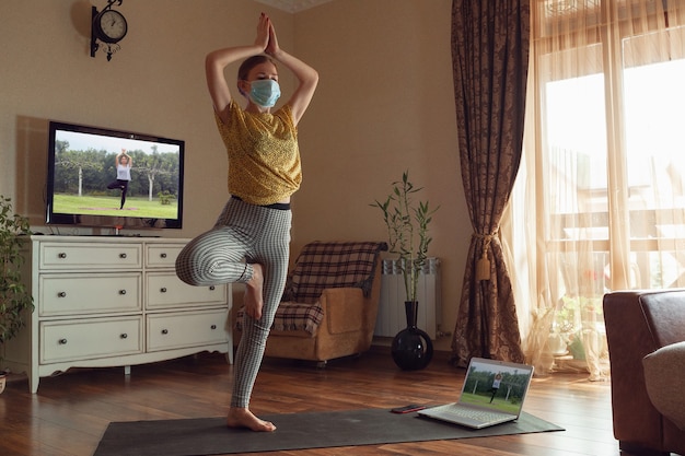 Sportieve jonge vrouw die online yogalessen volgt en thuis oefent terwijl ze in quarantaine is. Concept van een gezonde levensstijl, wellness, veilig zijn tijdens een pandemie van het coronavirus, op zoek naar een nieuwe hobby.