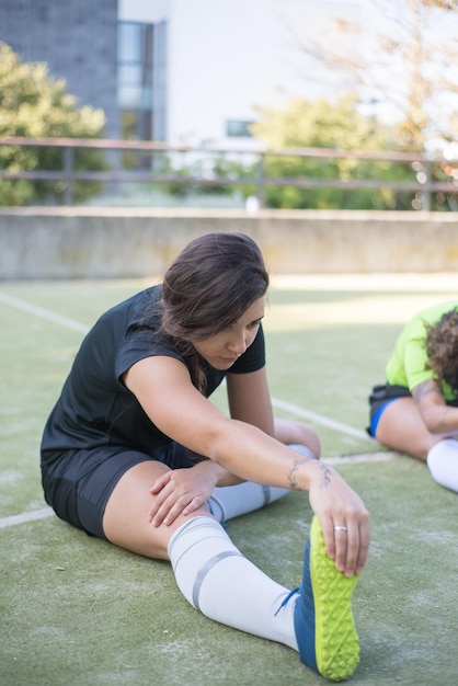 Sportieve jonge vrouw opwarmen op zomerdag. Geconcentreerde Sportvrouw in zwart uniform zittend op gras, stretching. Sport, vrije tijd, vrouwelijk voetbalconcept