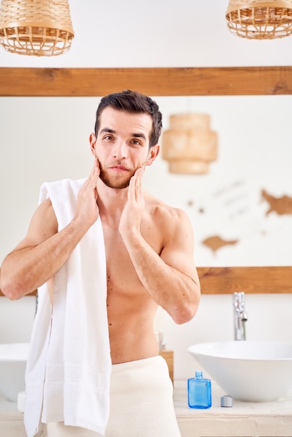 Sportieve jongeman wast zijn nek en staat met handdoek op zijn schouders voor badkamerspiegel bathroom