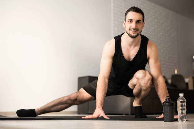 Sportieve man doet side lunge oefening. Mannelijke atleet met fit lichaam opwarmen, been stretch training thuis doen. Zorg voor jezelf! Moderne lichte kamer op de achtergrond. Zwarte sportkleding.