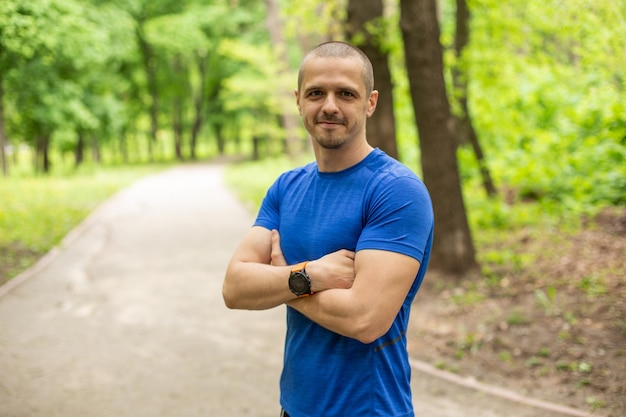 Sportieve man met gekruiste handen gekleed in een t-shirt die in de camera kijkt en in het stadspark staat