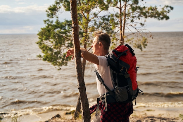 Sportieve man met rugzak staat op het strand en leunt tegen een boom terwijl hij geniet van het uitzicht