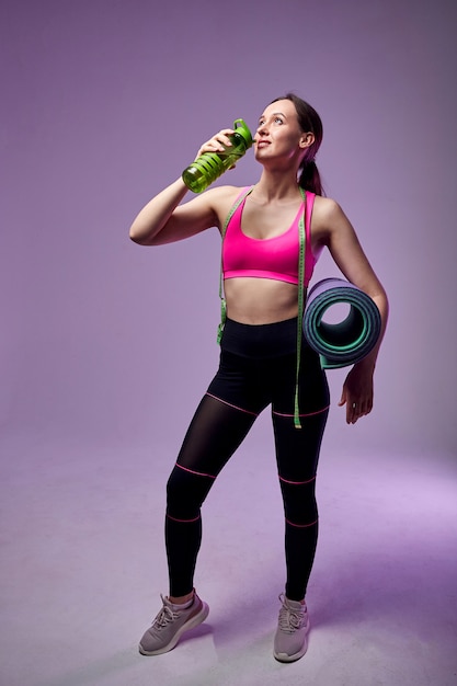 Sportieve mooie vrouw in sportkleding met een fles water en fitness mat geïsoleerd op violet. Naar de sportschool gaan.