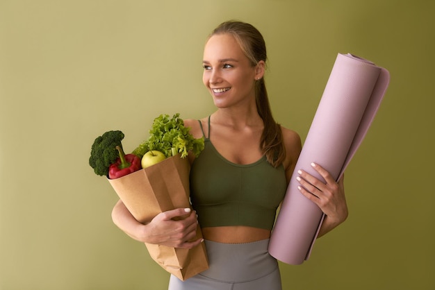 Sportieve mooie vrouw met een zak met groenten en een mat op een groene achtergrond Het concept van sport en gezond eten