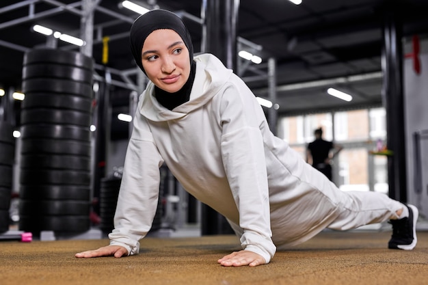 Sportieve Moslimvrouw doet push ups, fitness vrouw plank oefening uit te werken. gemotiveerde vrouw in hijab geniet van training op verdieping in sportschool